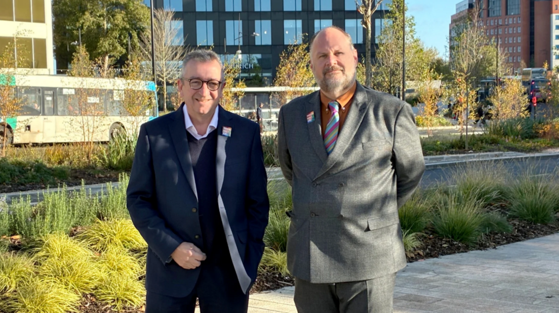 Two men stood side by side in an outdoor setting. One is in a suit jacket and smiling with glasses, the other is wearing a suit and colourful tie and orange shirt. A bus is passing in the background.
