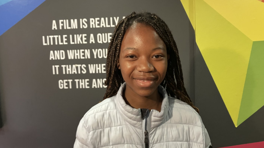 Shows a young black girl with breaded hair and wearing a grey zip-up puff jacket smiling and standing in front a partially-eclipsed sign about film.