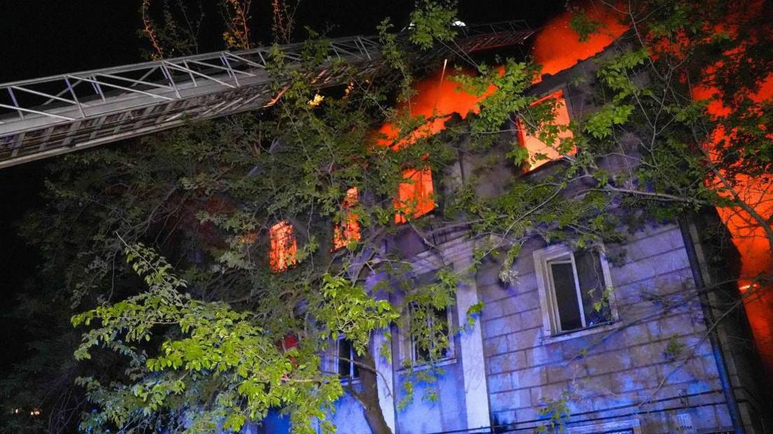 A residential building burns as a ladder extends from a fire truck that's out of view, with trees and foliage in the foreground, in Odesa on 1 May.