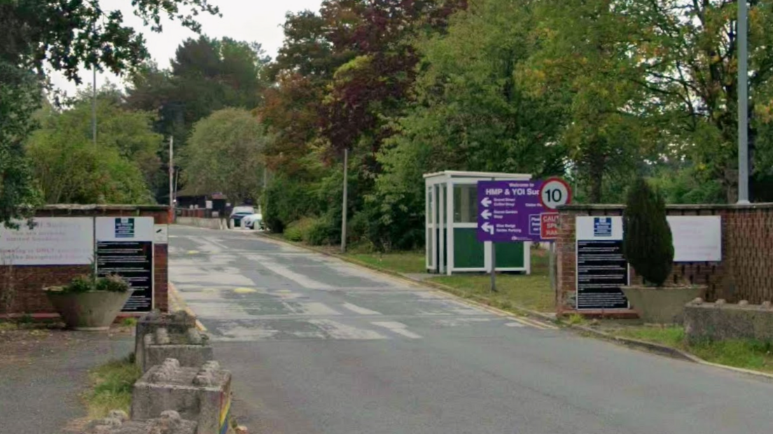 A road runs between two walls, with a number of signs on both walls showing that it is a prison. A larger sign behind the wall on the right says HMP and YOI Sudbury