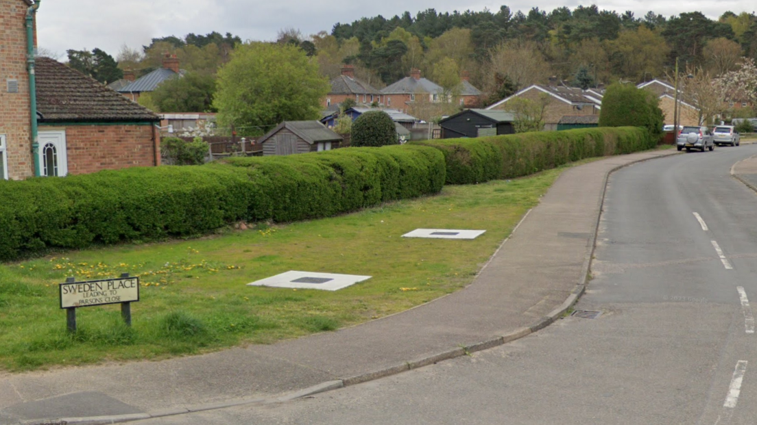 A residential road with a path and hedges. There is a road sign saying "Sweden Place" in the bottom left corner and houses can been seen in the background and cars are parked in the distance. 