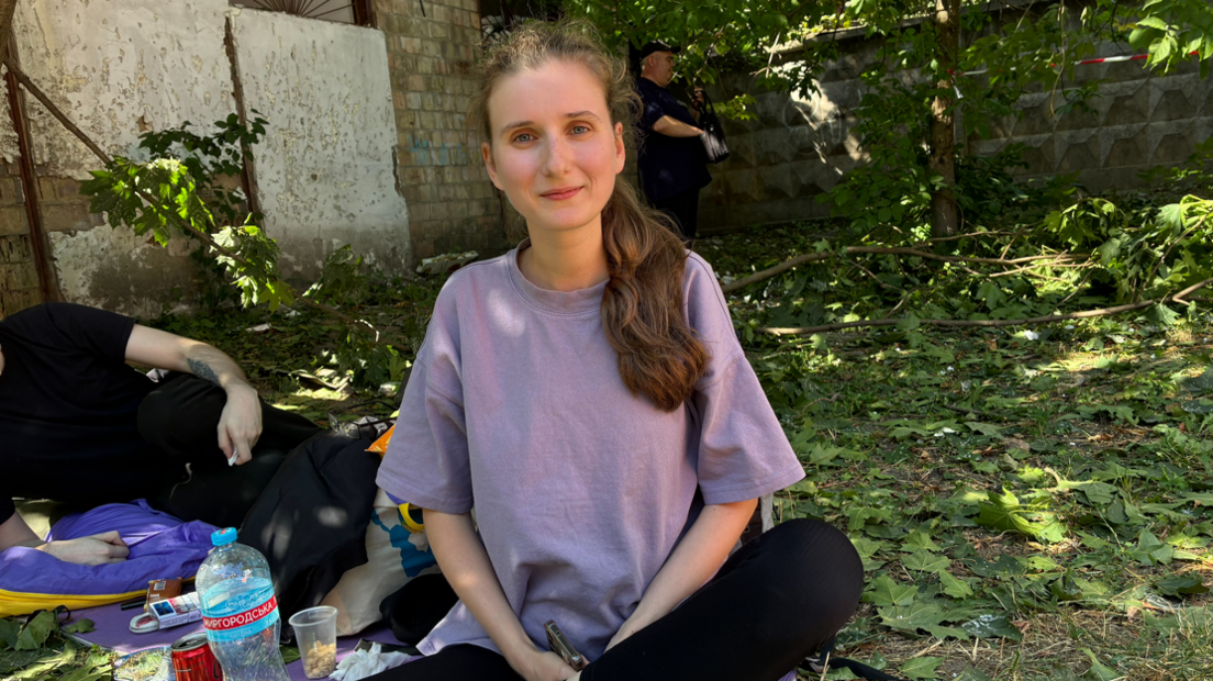 Anastasiia Kovtun sits crossed legged on the ground with leaves around her and a man sat behind her