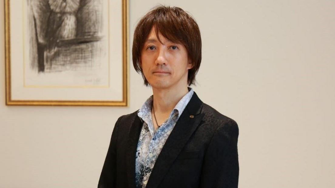 Tomohiko Sho stands in front of a wall with a picture of an owl. Mr Sho is wearing a blazer over a colourful blue shirt with an open top button. He is Japanese. He has a neutral expression and smart medium-length hair.