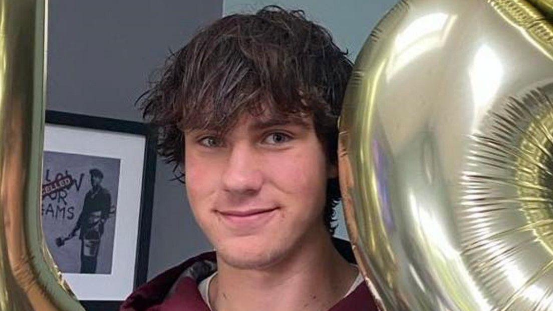 A man with dark hair is looking at the camera. A grey background is behind him on the left.