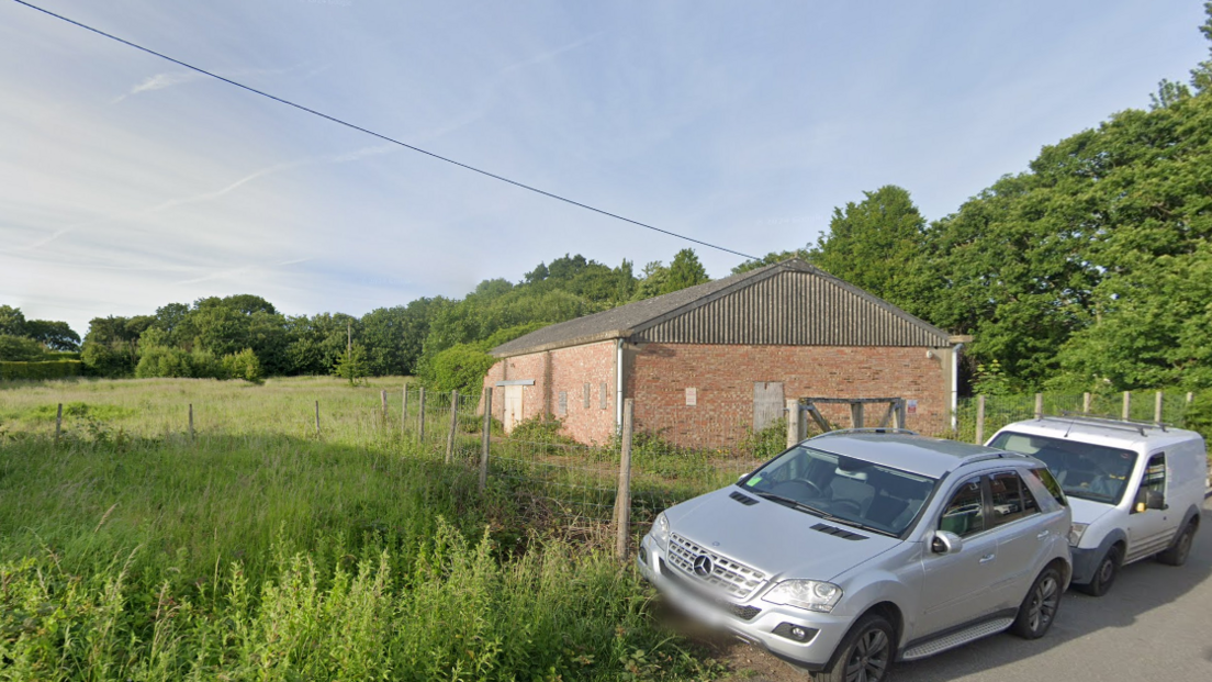 A field with an outbuilding in the foreground. The building has two cars parked on a verge next to it.