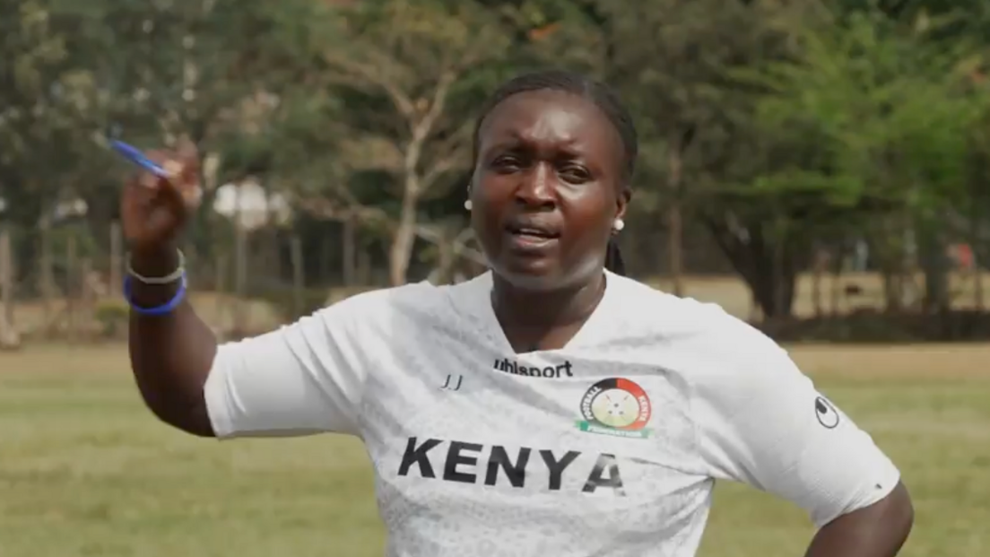 Jackline Juma, wearing a white training shirt with the word Kenya on it, gestures with her right arm as she holds a pen in her hand