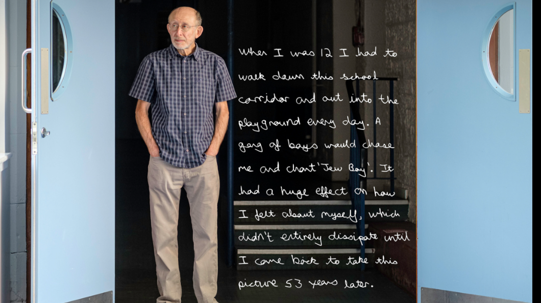 Jeremy, a senior man, is stood at the entrance to a school corridor in Trinity Academy, Edinburgh. He stands with his hands in his pockets, and his expression appears both reflective and sad.