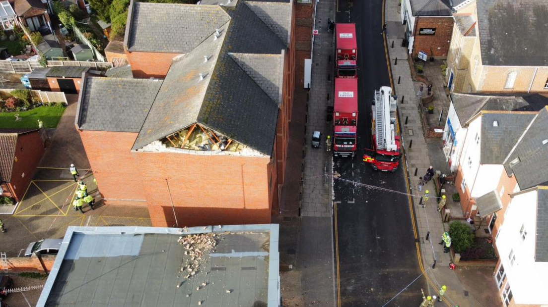 Drone image of a fire engine and two red lorries on an otherwise vehicle-free High Street. Several fire fighters and police officers are standing around. In the centre of the image a red-brick building has its gable end of bricks missing. Rubble can be seen on a neighbouring flat roof at a lower level next to it.