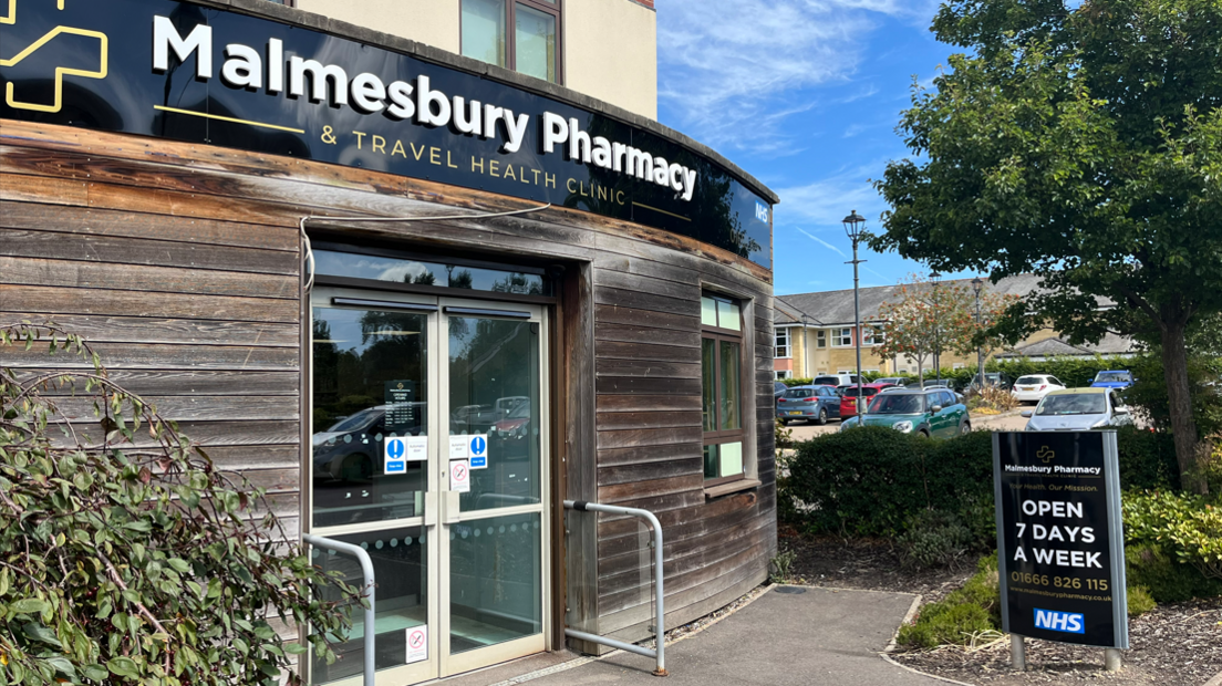 An entrance to a new pharmacy. It has wooden cladding on the outside and the words "Malmesbury Pharmacy" on a large sign, in white letters on a black background