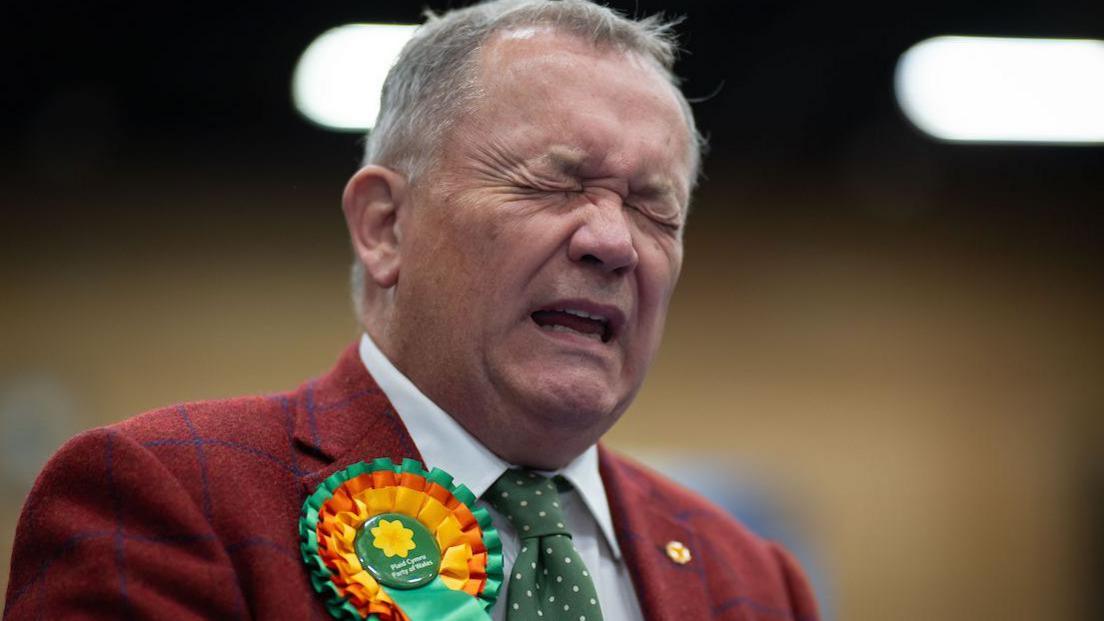 A man in a red jacket is seen closing his eyes, looking tense