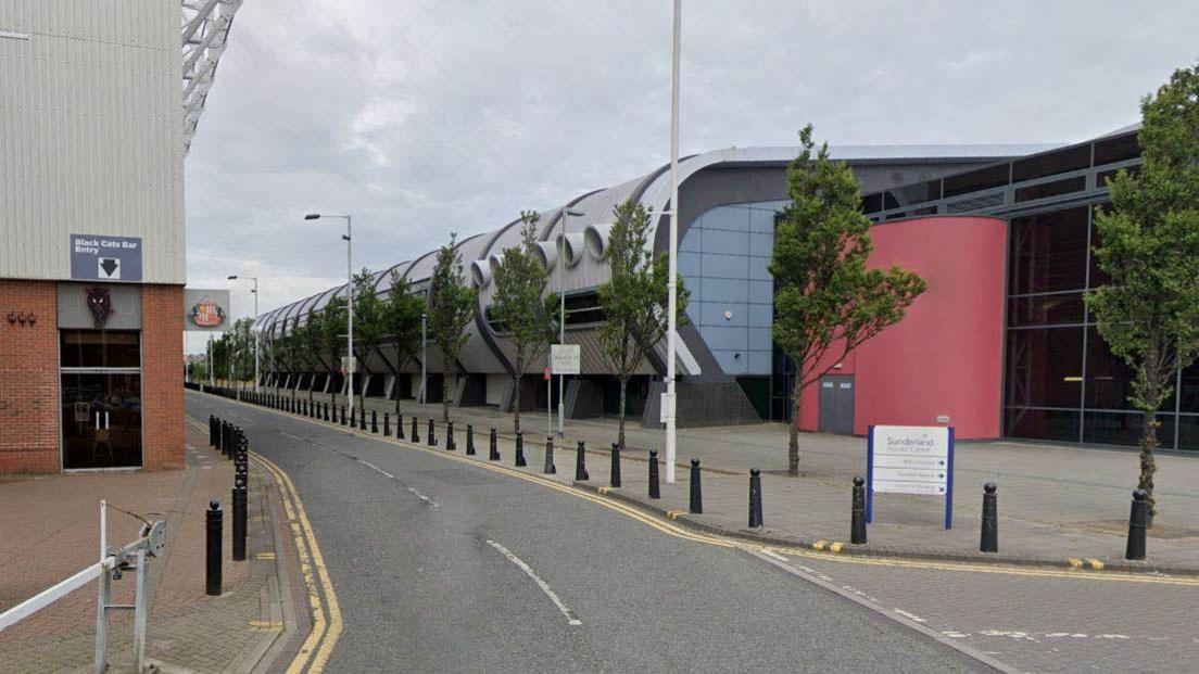 Sunderland Aquatic Centre stands to the right of the Stadium of Light. The Aquatic Centre has a large curved wall which along the length of the road. It is grey with a red wall near its entrance. The side of the Stadium of Light can be seen to the left. A sign with the club's badge is hanging from the wall while a door is marked with a black cat emblem.
