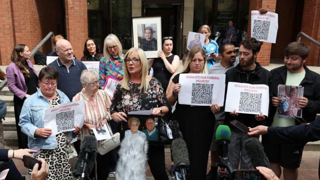  “REGULATE OUR FUNERAL INDUSTRY” with large QR codes, while others hold framed and printed photographs of older adults. In the centre, a person speaks while holding a framed photograph, with supporters gathered closely around. The building entrance and steps form the backdrop.