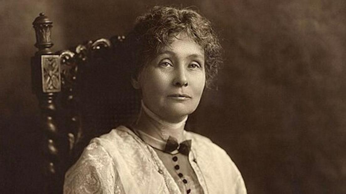 A dark sepia toned image of Emmeline Pankhurst sat in an ornate wooden chair. She has dark hair tied up with wavy tendrils falling around her face. She has a lacy shawl over her shoulders.