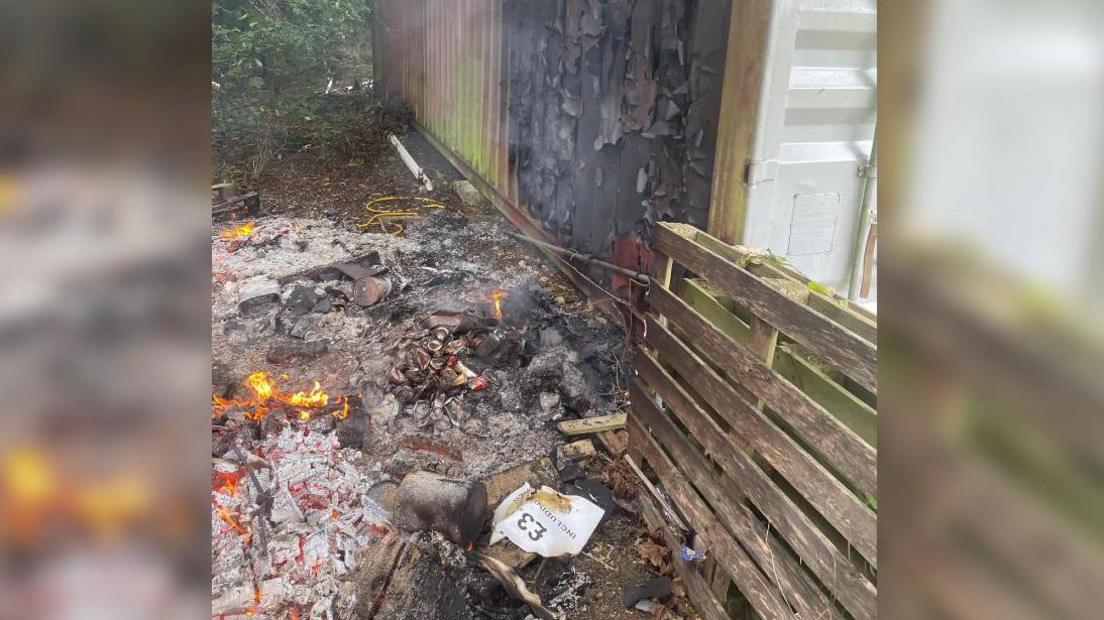 The exterior of a storage container. The paint on the metal corrugated walls has been blackened and is peeling off. On the ground there is a pile of grey ashes still on fire and a wooden pallet leading up against the container. 