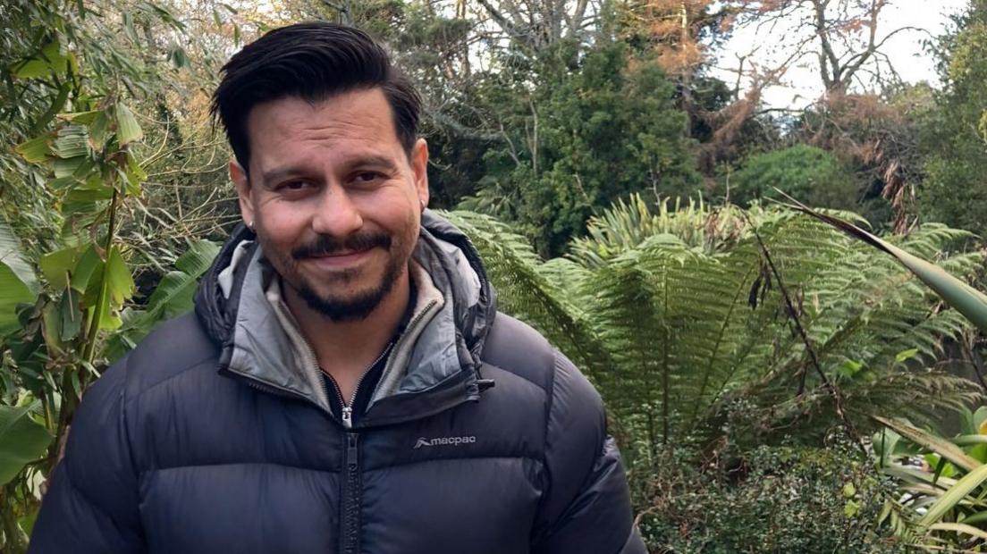 José Palma smiles at the camera, he is wearing a black puffer jacket and has a goatee and moustache. There are plants behind him.
