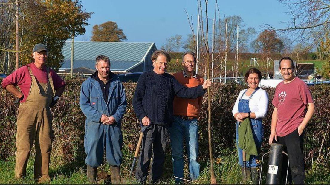Six volunteers pose with gardening tools whilst undergoing a tree planting event. Behind them is a hedgrow and some buildings.