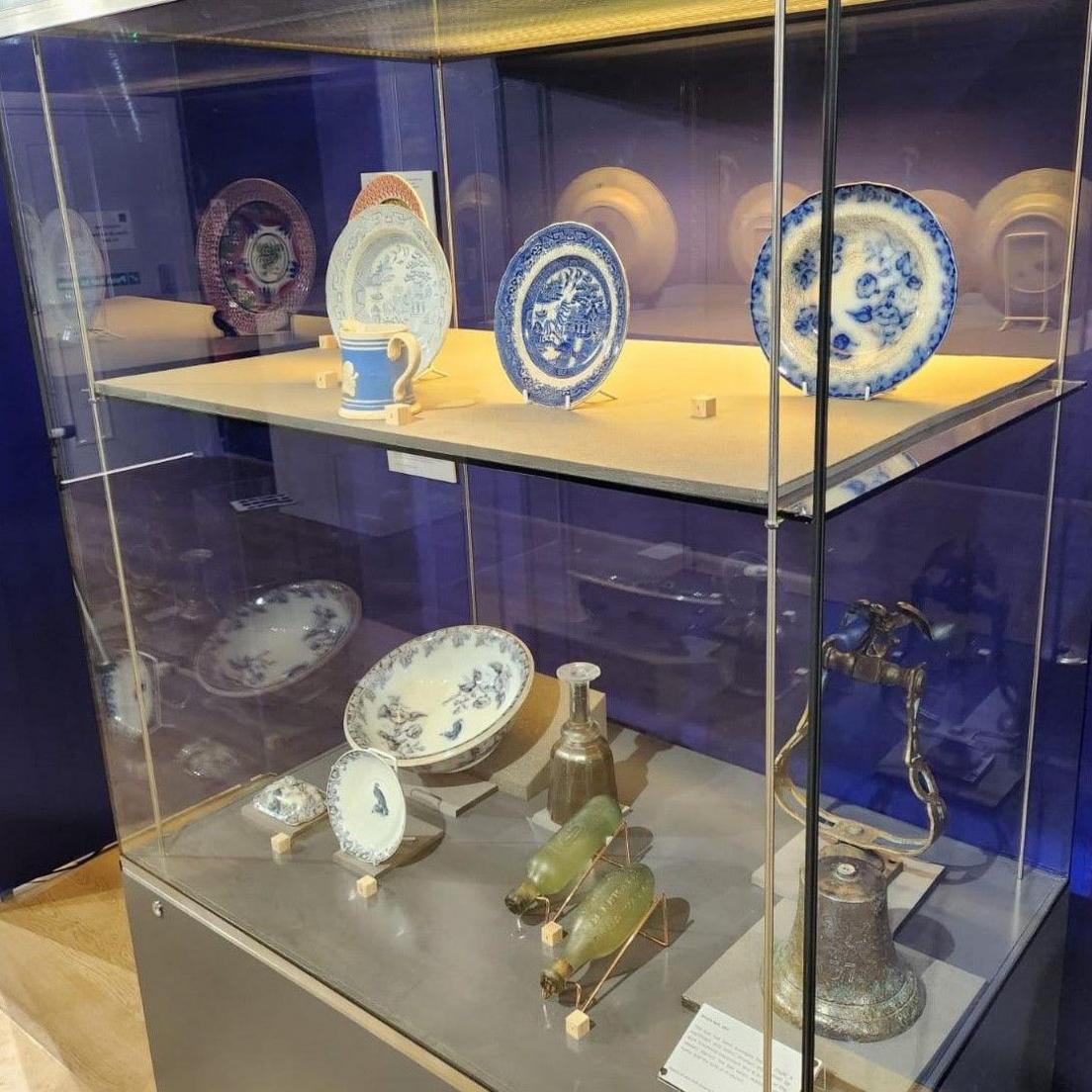 A two-shelf glass display cabinet with four plates and a cup in blue and white on the top level, and a mix of items on the bottom including bowls, a small bell and a pair of small green glass bottles.