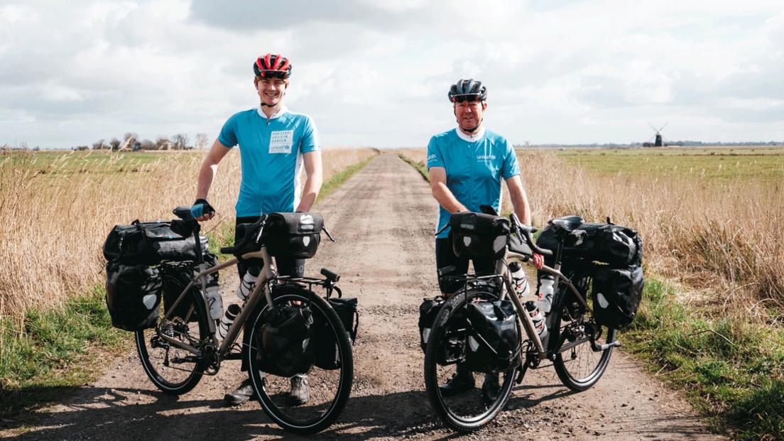 Joshua and George Kohler, both wearing cycle helmets, pale blue cycling tops and black cycling trousers. They are standing by and holding their bikes, which are laden with black panniers, and are pictured on a track in Norfolk, with flat fields either side and a windmill in the distance