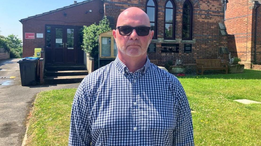 The image shows a man standing outdoors in front of a red-brick church. He is wearing dark sunglasses and a long-sleeved, button-up shirt with a blue-and-white checkered pattern. The man has a shaved head and is standing upright on a grassy area.