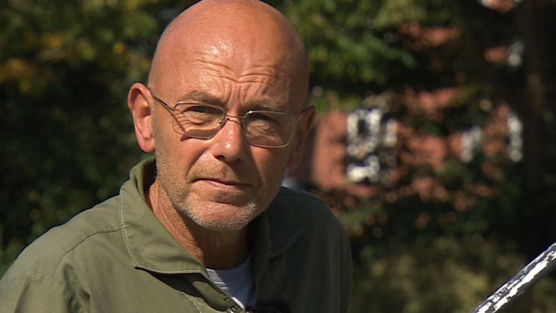 A head and shoulders shot of Wayne Hemingway. He is slightly to the left of frame and is wearing an olive green shirt with a white T-shift underneath. There is soft-focused vegetation in the background