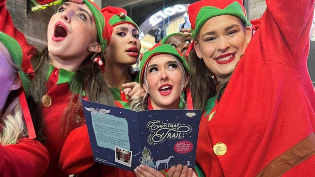 Five young women dressed as elves with one holding a Christmas Trail leaflet in a shopping centre. They all have red lipstick on and are smiling.