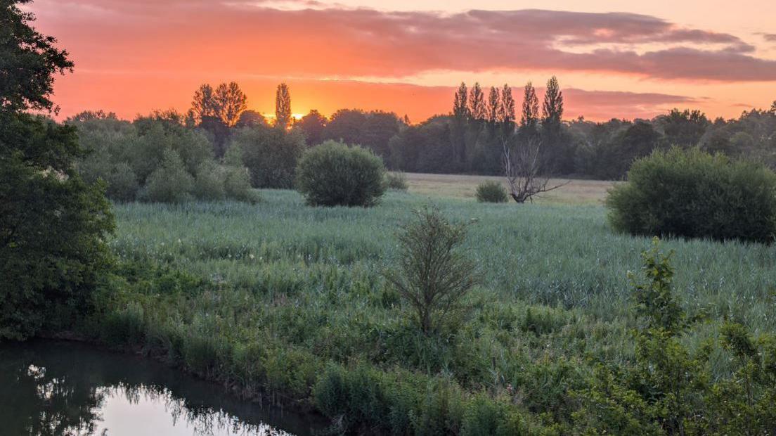 A field in Oxford next to water and being overlooked by the vibrant orange colours being provided by either a sunset or sunrise 