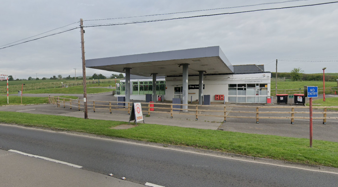 A small flat-roofed building with what looks like an old petrol station forecourt. The sky is grey, and green fields surround the building, with an A road in front of it.