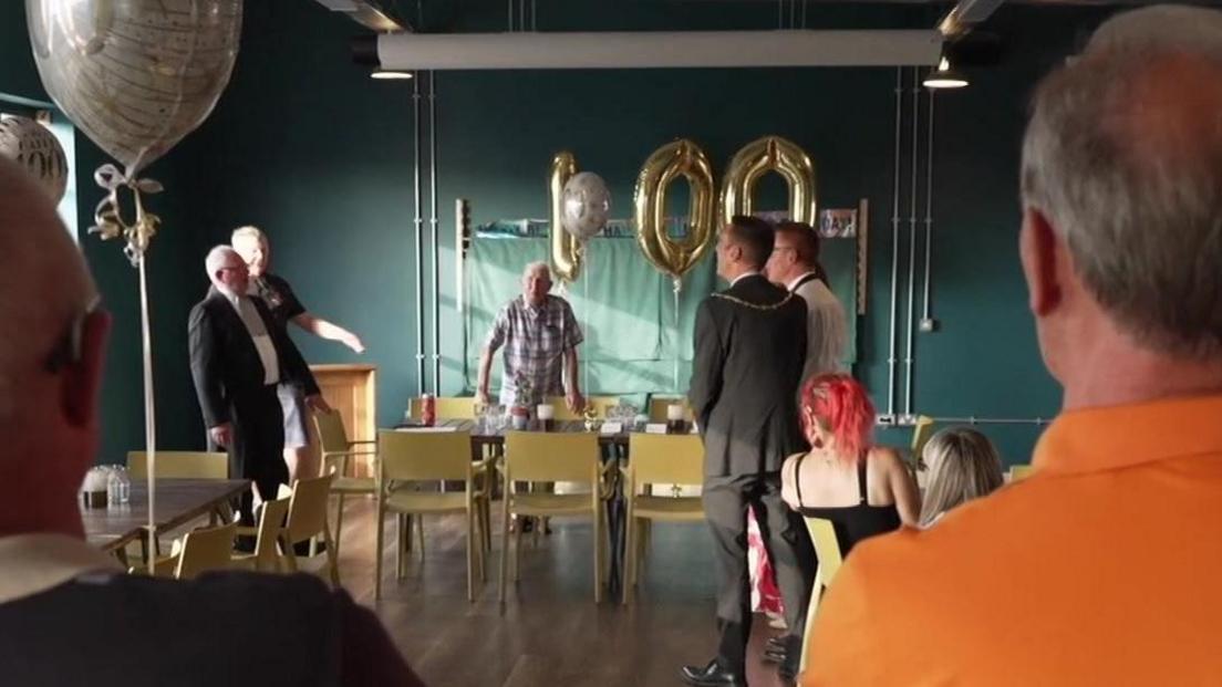 An old man wearing a check shirt stands behind a table and chairs. Above him gold balloons spell out the number one hundred.