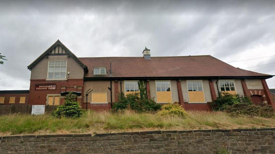 A large red-brick building, some of its windows are boarded up, a sign says Brierley Village Club.