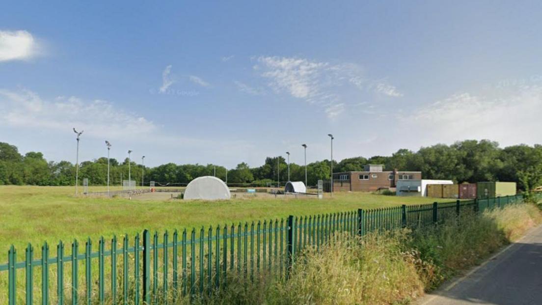 A green field behind a dark green iron fence. A brown brick building and a old cricket pitch and floodlights are in the middle of the picture. 