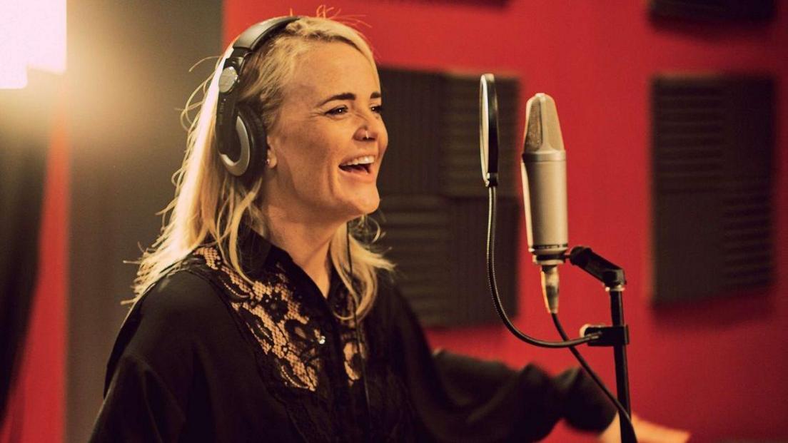 Bristol singer Elles Bailey smiles as she rehearses in a recording studio. She is wearing a black top and has blonde hair