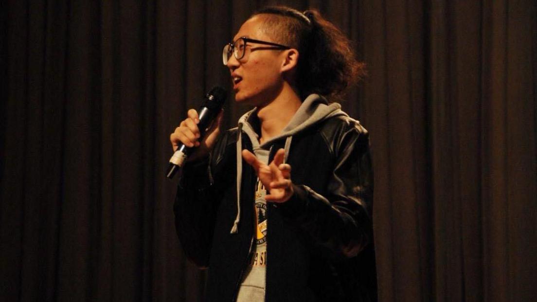 Chizi performs on stage in 2016, wearing a grey hoodie under a black jacket, with his long hair tied back.