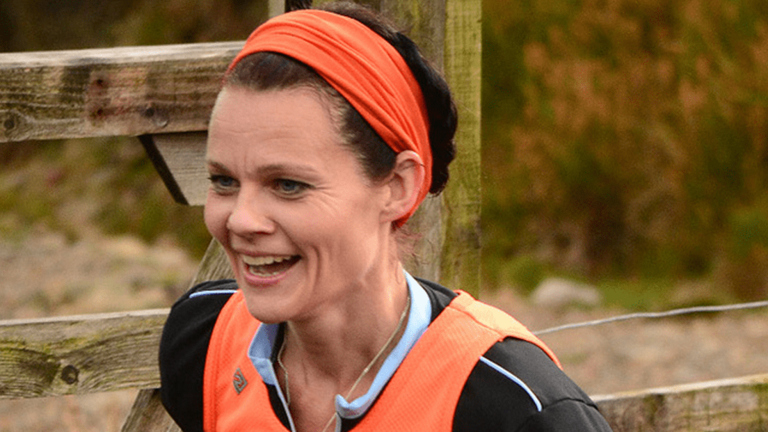 Emma Gregory running on fells, wearing an orange headband and orange running vest, with a black tshirt. She has her dark hair tied back and is smiling as she is mid-stride.