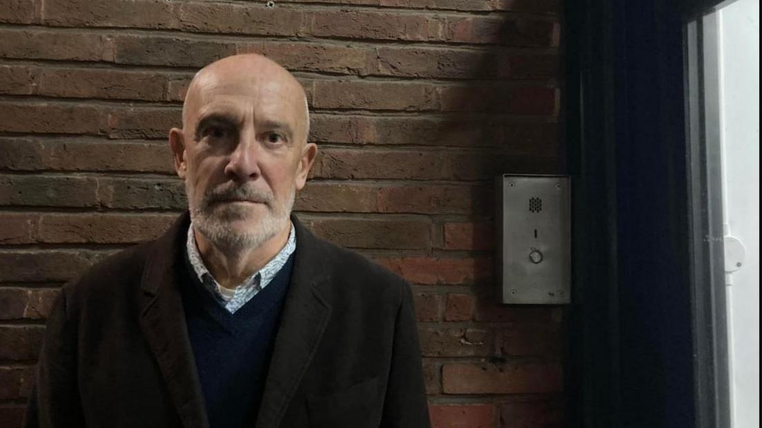 A bald man with a grey beard, wearing pale blue shirt, dark blue jumper and brown coat stares into the camera.  