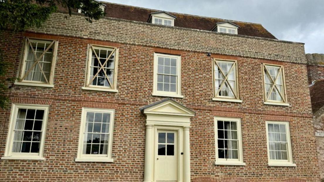 The cream and red Georgian facade of Burstow Farmhouse, with five windows along the first floor and four windows on the ground floor, two either side of the neo-palladian front door.