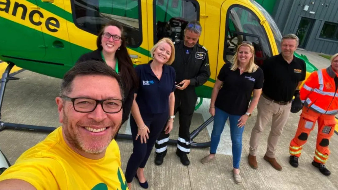 Mike Land in a bright yellow t-shirt taking a selfie with several members of the Wiltshire Air Ambulance, all in uniform, and standing in front of an air ambulance