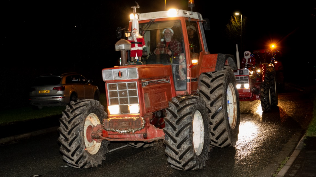 A red tractor in the dark with a Santa figure on the front and the driver wearing a Santa hat. Another red tractor is following close behind