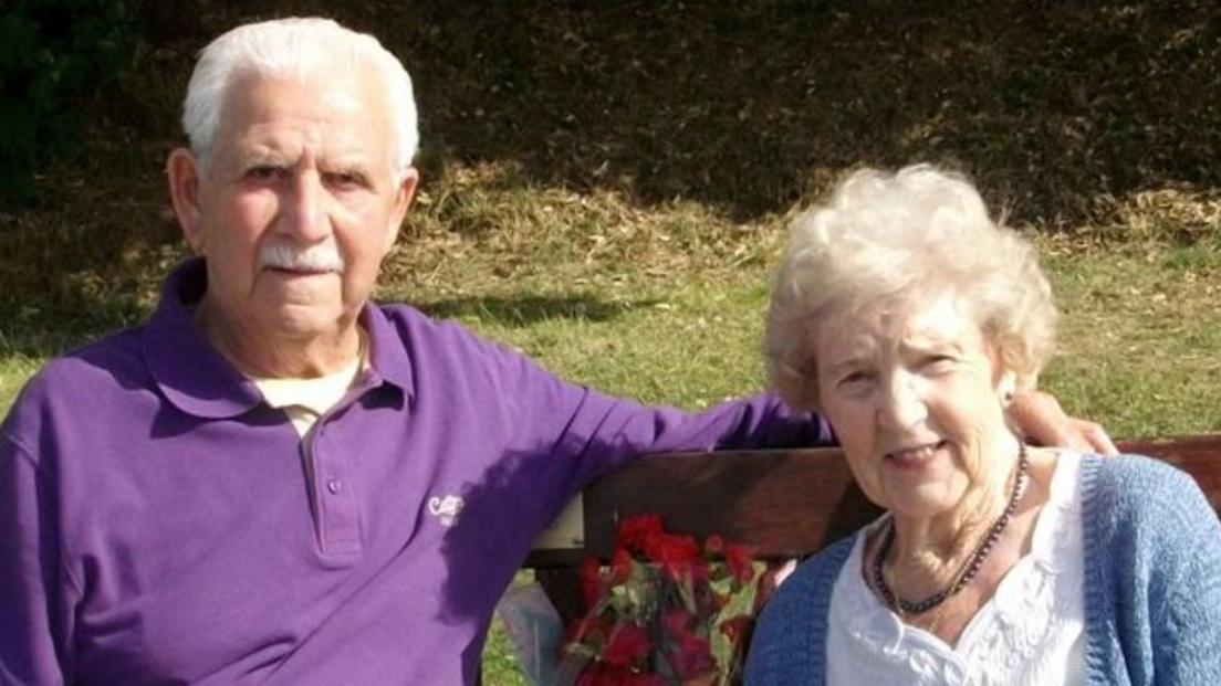 Bill and Lucy Warren sit on a memorial bench for their son Peter. A bunch of roses are between the couple. Bill has white hair and a white moustache. He is wearing a purple Cotton Traders polo shirt. Lucy has white hair and is wearing a dark necklace, white shirt and blue cardigan.