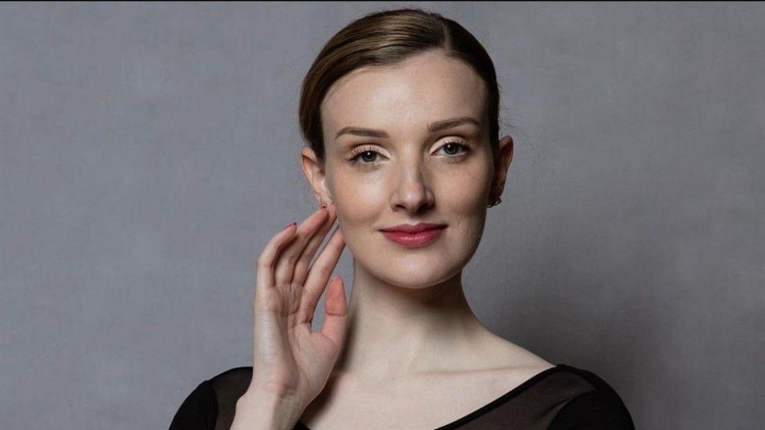 A woman is shown from the shoulders up, smiling with her mouth closed and looking at the camera. Her hand is raised and touching the side of her face. She has brown hair tied up and slicked back. She is wearing a black top. She is in front of a grey background.