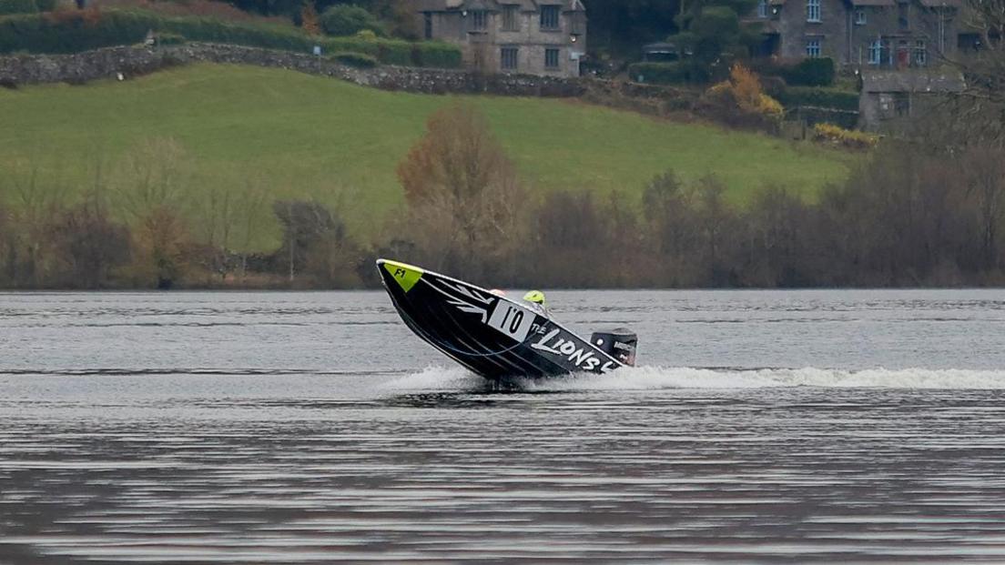 Dark blue boat in the lake with its nose pointing into the air. There are hills in the background and a couple of large houses. The boat has the number 10 written on its side.