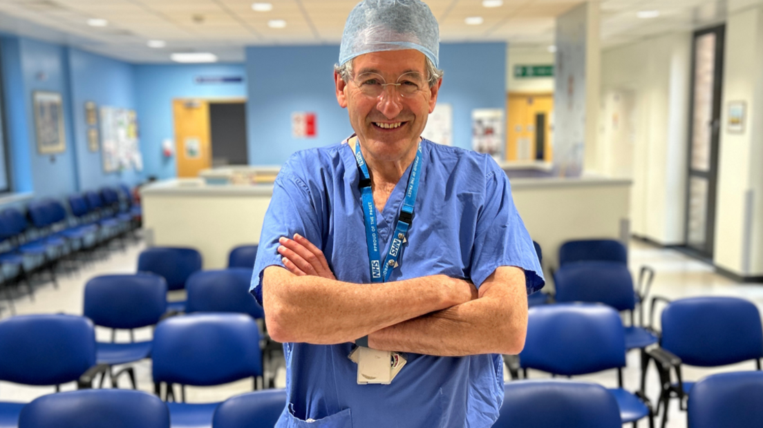 Peter Prinsley MP is smiling at the camera in a hospital waiting room. He is wearing a surgeon's blue scrubs and hat