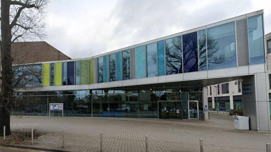 The outside of a two-floor building, mostly made of glass. Some of the upper panels are blue or yellow. In front of the building is a large public walkway.