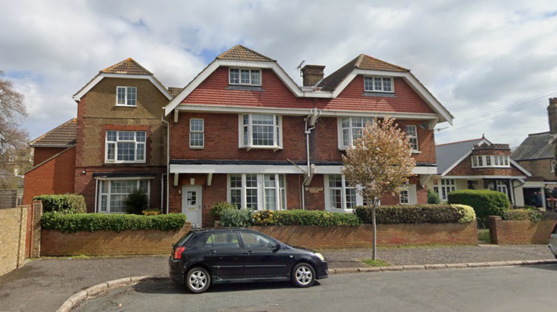 A large three-storey building made of red and yellow bricks, with a roof pointed in three places and large, white windows. A small black car is parked on the road in front of the property.
