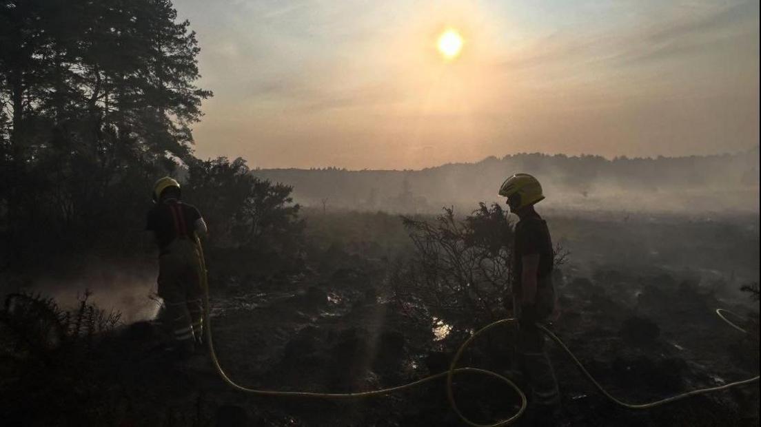 Two firefighters at dusk holding a yellow hose on heathland where smoke is seen rising.