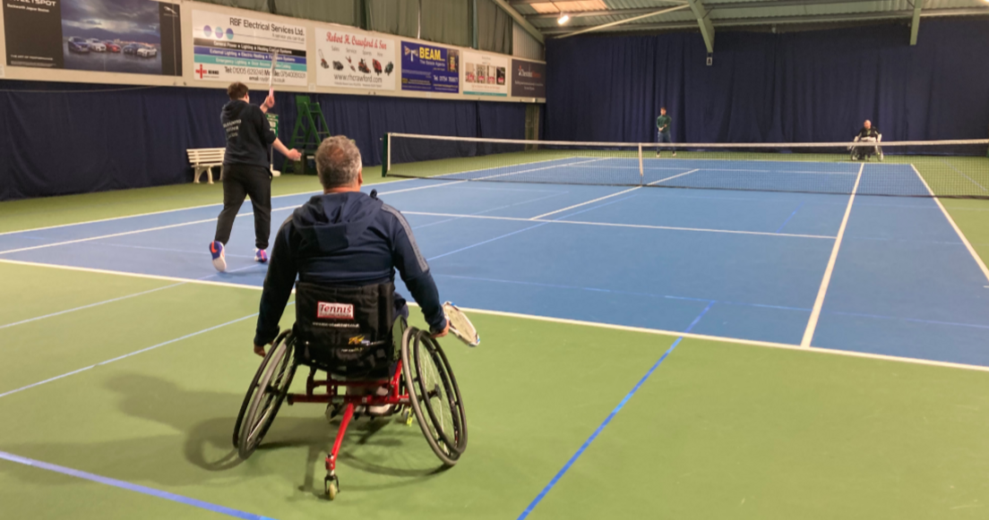 The picture is taken from the back of an indoor court. The floor of the court is blue and green. The two wheelchair players are both at the back of the court behind the serving line, and the able-bodied players are at the front.