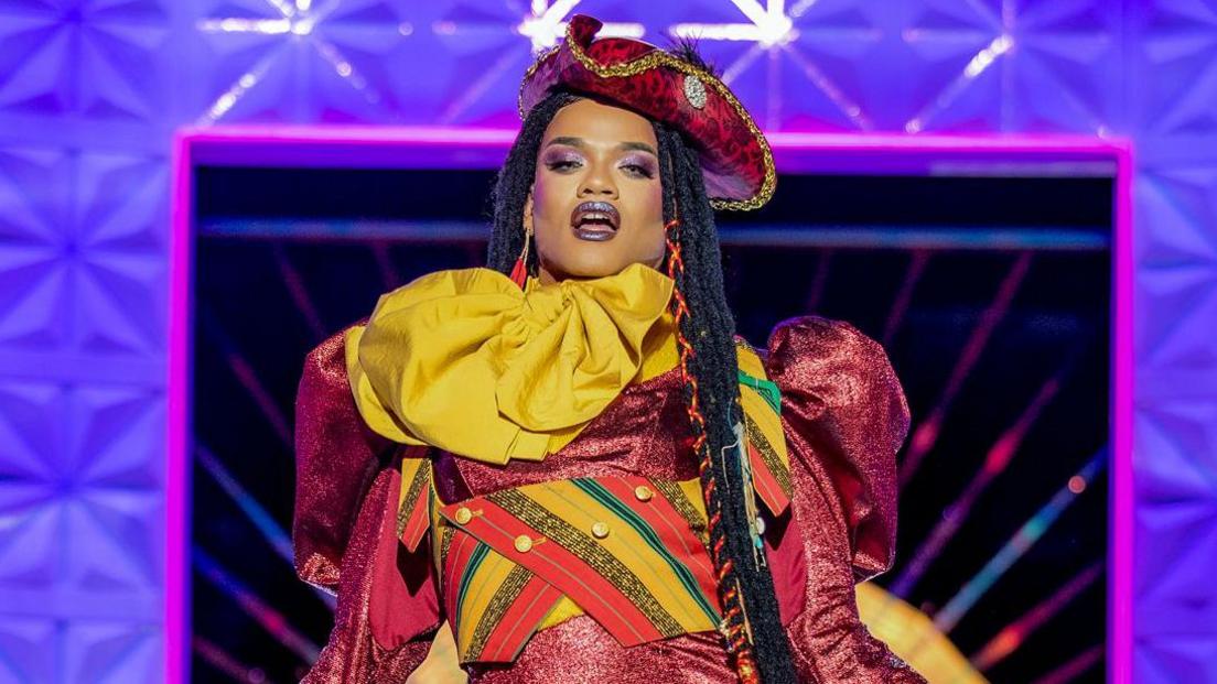 Rileasa Slaves on the runway. She wears a red pirate-style hat, long braids and a red puff-sleeved dress matched with yellow scarf and a cropped waist coat in red, yellow, green and gold stripes.