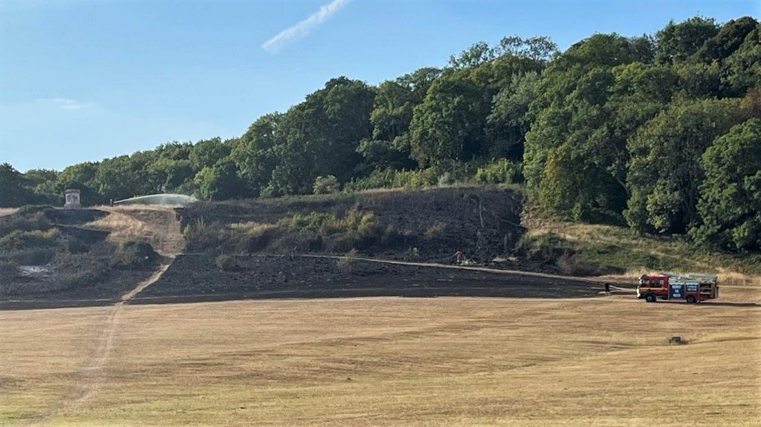 A large area of burnt grass on Stoke Park Estate. There is a fire engine parked in the bottom left corner and a large hose at the top of the hill which is dampening down the grass.
