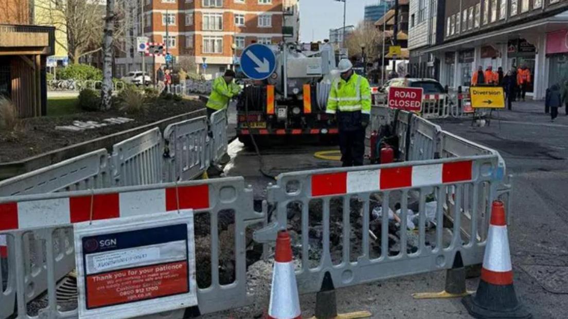 SGN gas workers in hard hats and hi vis inside a coned off area in a busy street in Woking with plastic barriers and pumping equipment in background