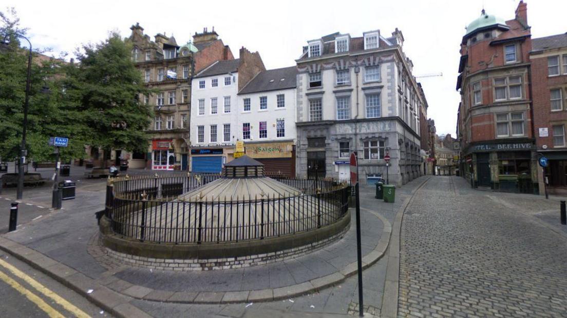 Streetview of a cobbled square with three and four storey buildings housing pubs and nightclubs. In the foreground is a round domed roof protruding from the square.