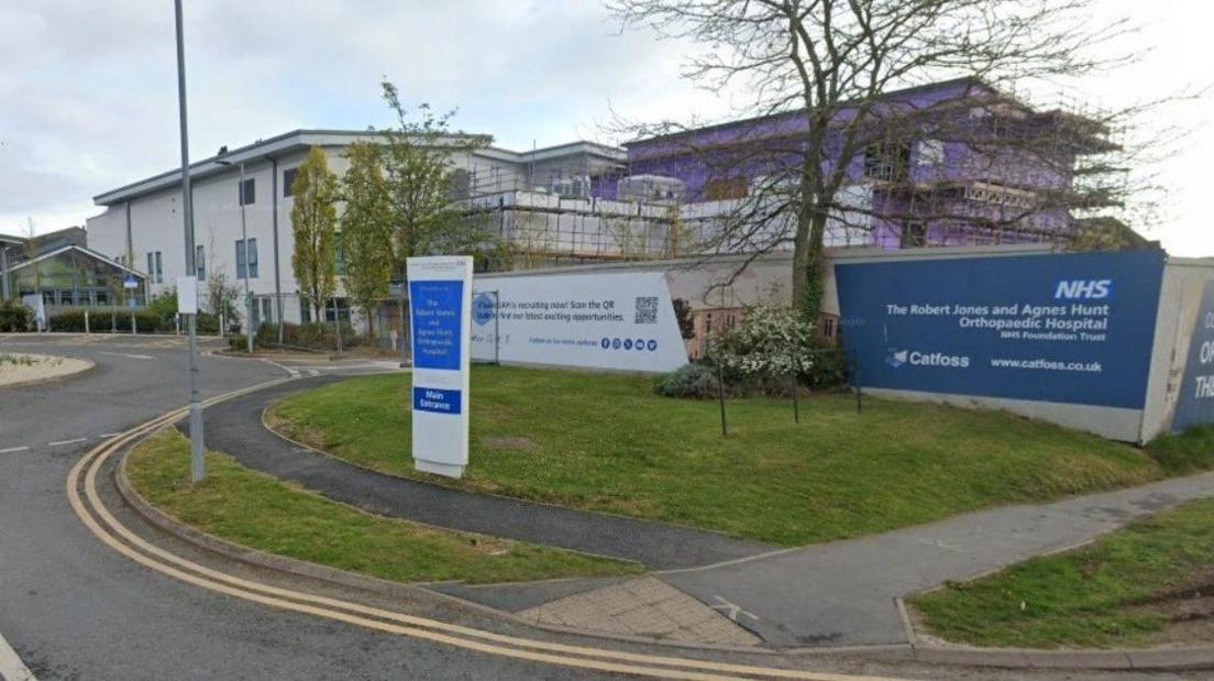 A blue sign with the trust's name in white writing is on the right of the image. A tree and a purple coloured building are behind it on the photo and another building is prominent on the left of the image.